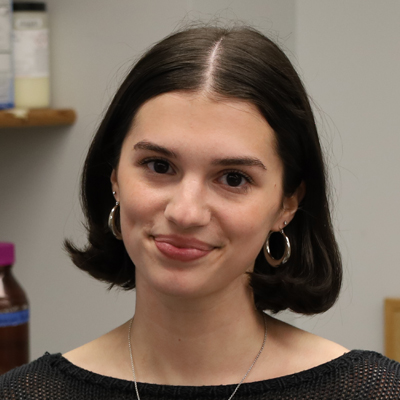 The image shows a person with short, dark hair wearing a black top and a necklace. The background includes a shelf with various bottles and containers.