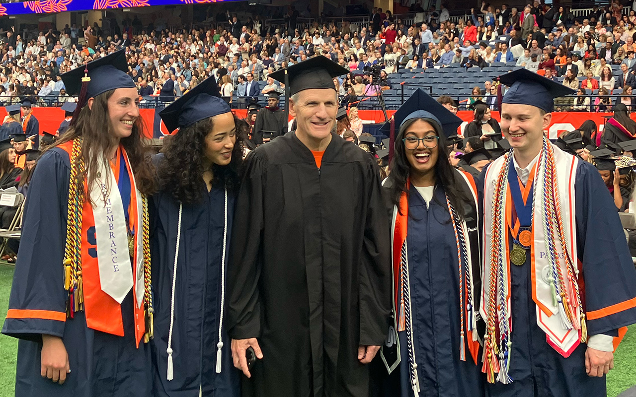 A group of five people posing for a photo at a graduation ceremony. Four individuals are wearing blue graduation gowns and caps, adorned with cords, stoles and medals. The person in the center is dressed in a black gown and cap. Behind them, an audience is seated in bleachers.