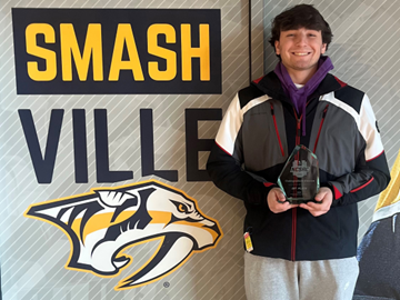 Individual standing in front of a wall with the text 'SMASH VILLE' and a saber-toothed tiger head logo, holding an award.