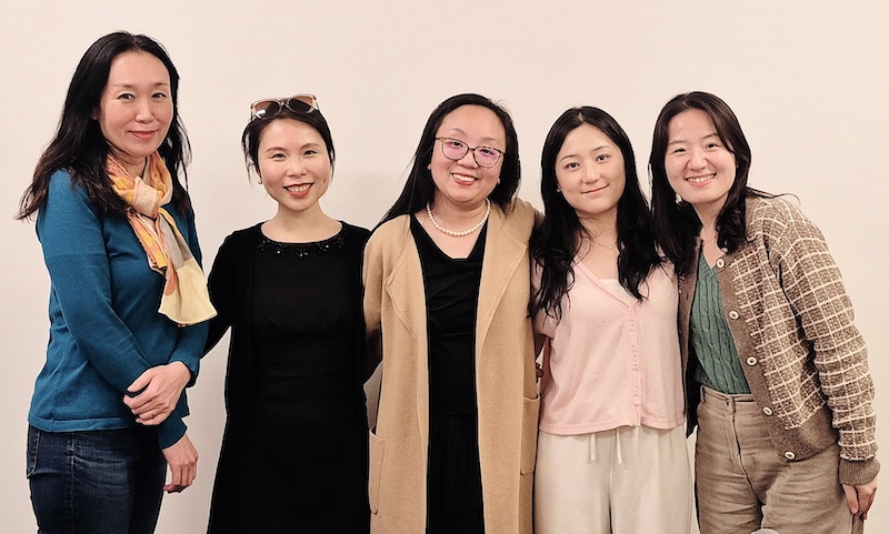 Five people standing side by side against a plain background. From left to right: the first person wears a blue long-sleeve shirt, jeans, and a light-colored scarf; the second person is in a black outfit with sunglasses on their head; the third person wears a beige cardigan over a black top and pants, accessorized with a necklace; the fourth person is in a light pink short-sleeve shirt and white pants; and the fifth person wears a green top under an open brown plaid jacket with beige pants.