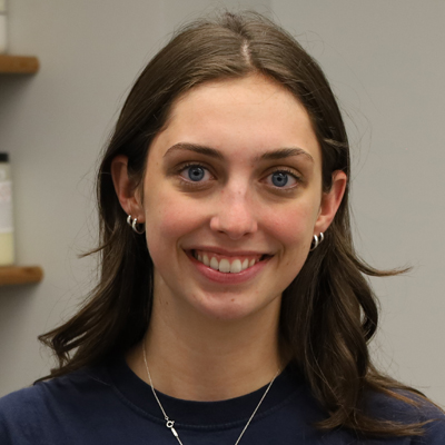 The image shows a person with shoulder-length brown hair wearing a navy blue shirt and a silver necklace with a small circular pendant. The background includes shelves with various items, including a jar on the left side.