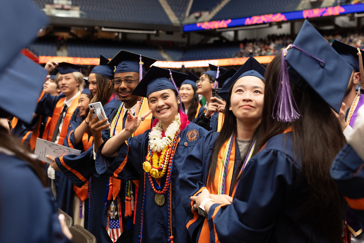 A group of graduates wearing caps and gowns at Commencement 2025. The graduates are holding up their phones, possibly taking photos or videos. Some of the graduates are adorned with leis and honor cords. The background shows stadium seating with some spectators visible.