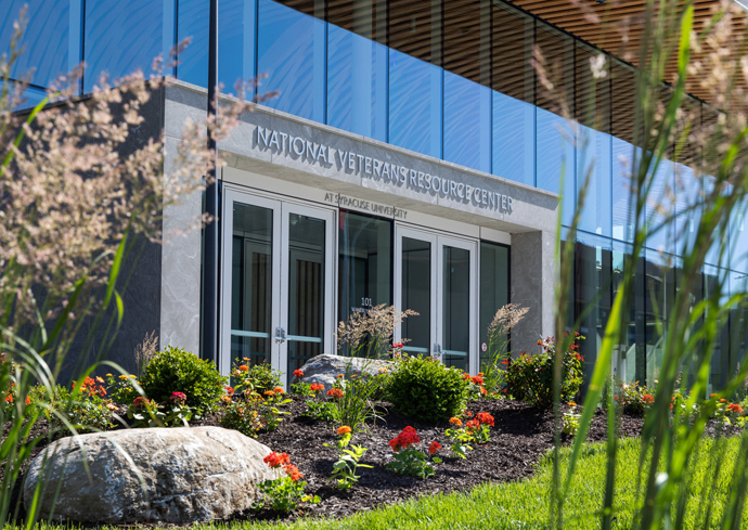 The entrance to the National Veterans Resource Center at Syracuse University. The building features a modern design with large glass windows and a sign above the entrance that reads "NATIONAL VETERANS RESOURCE CENTER AT SYRACUSE UNIVERSITY." In front of the building, there is a well-maintained garden with various flowers and plants, as well as a large rock on the left side. The sky is clear, and sunlight illuminates the scene.