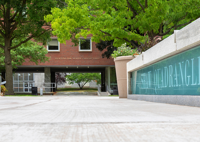 The image shows a view of a university campus area with a focus on the "Law Quadrangle" sign etched on glass in the foreground. In the background, there is a brick building with trees and greenery around it. The building has two floors visible, with windows and an entrance pathway leading through an archway under the building. The scene is bright and appears to be taken during daytime.