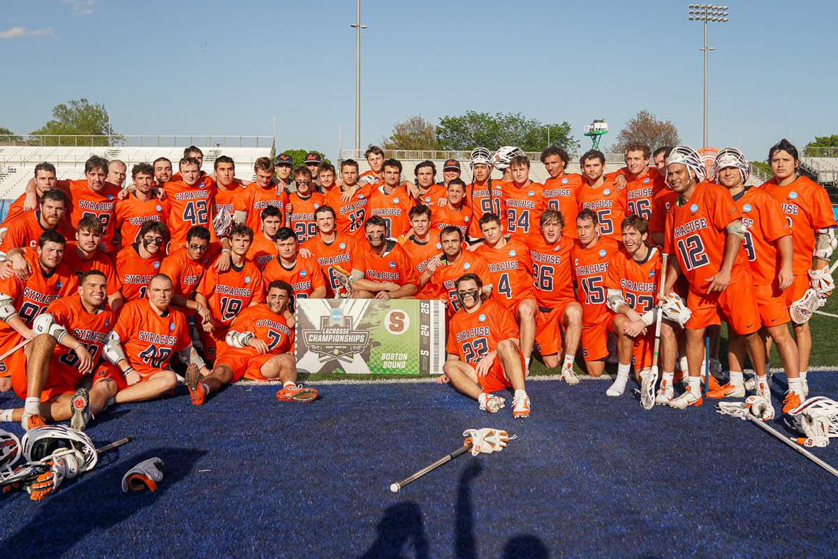 A large group of lacrosse players in orange uniforms and white helmets pose together on a field, holding lacrosse sticks. At the center, several players display a banner that reads NCAA lacrosse championships Boston bound. The background features a clear blue sky, trees, and stadium lights.