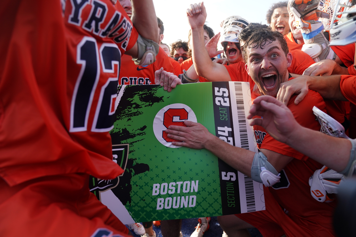 A group of people in orange jerseys celebrate while holding a large novelty ticket that reads 'Boston Bound.'