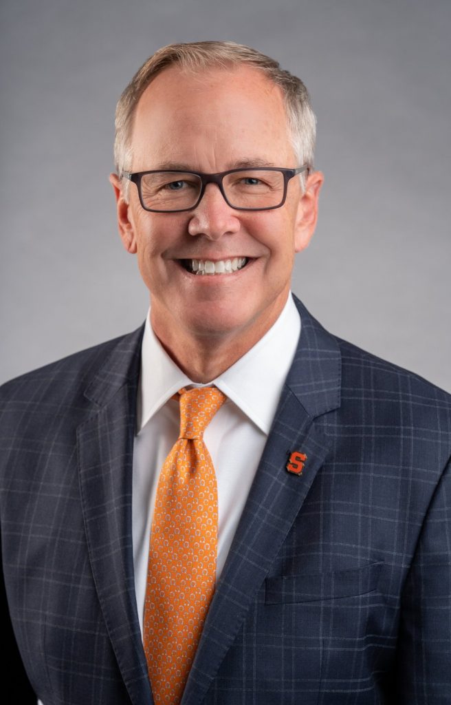 A person is wearing a dark blue checkered suit, a white dress shirt, and an orange tie with small white patterns. There is an "S" pin on the left lapel of the suit jacket.