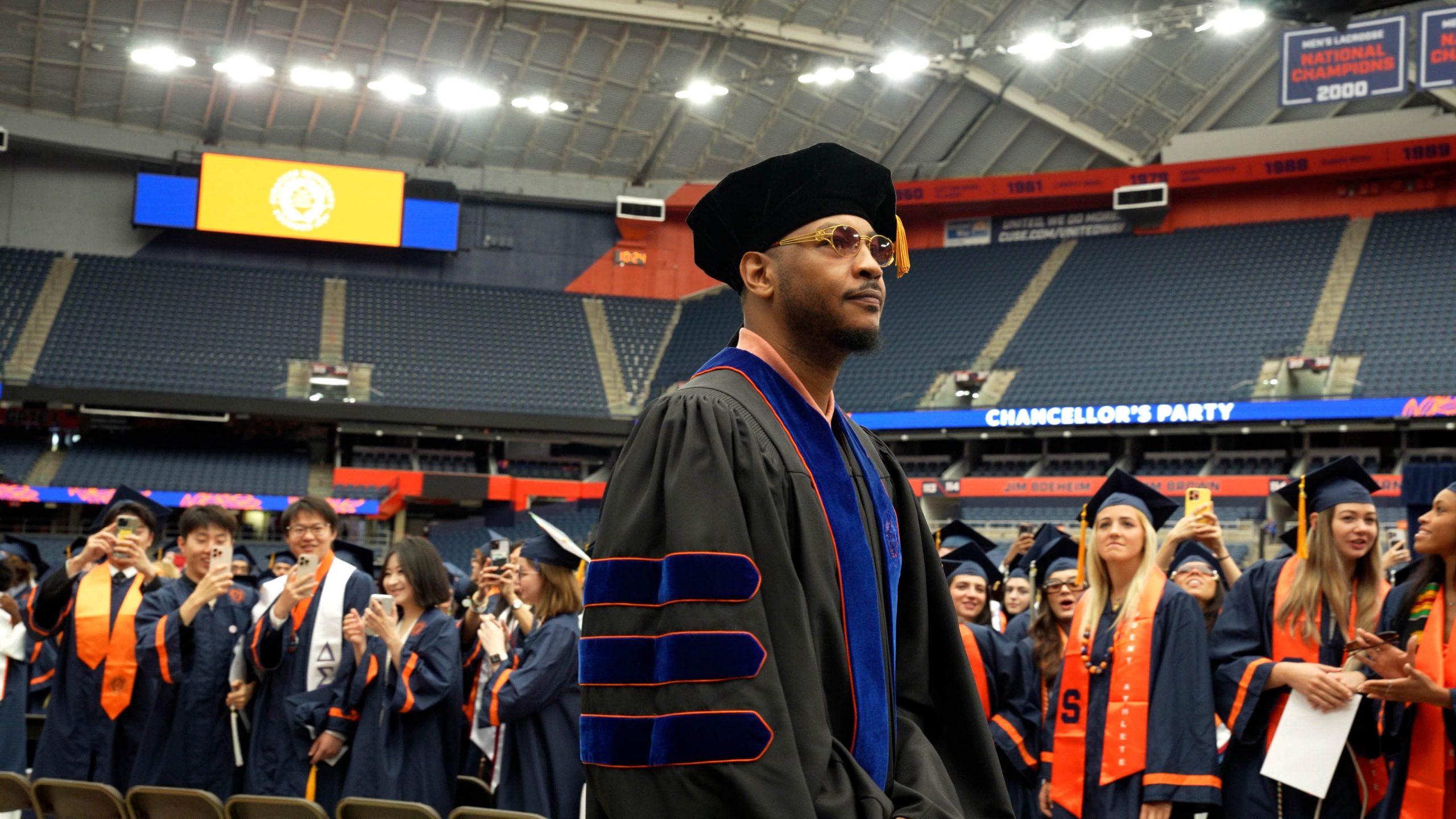 Carmelo Anthony wears academic regalia while processing at Commencement 2025. In the background, there are many other graduates dressed in similar attire, some of whom are taking photos or looking towards the stage.