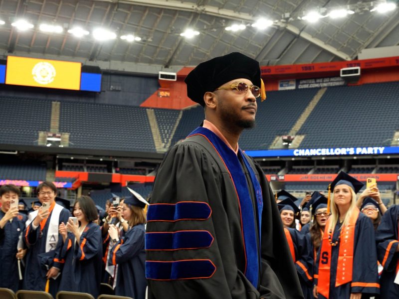 a graduation ceremony taking place in a large indoor stadium. The focus is on an individual in the foreground wearing academic regalia, including a black cap and gown with blue and orange accents. In the background, there are many other graduates dressed in similar attire, some of whom are taking photos or looking towards the stage.