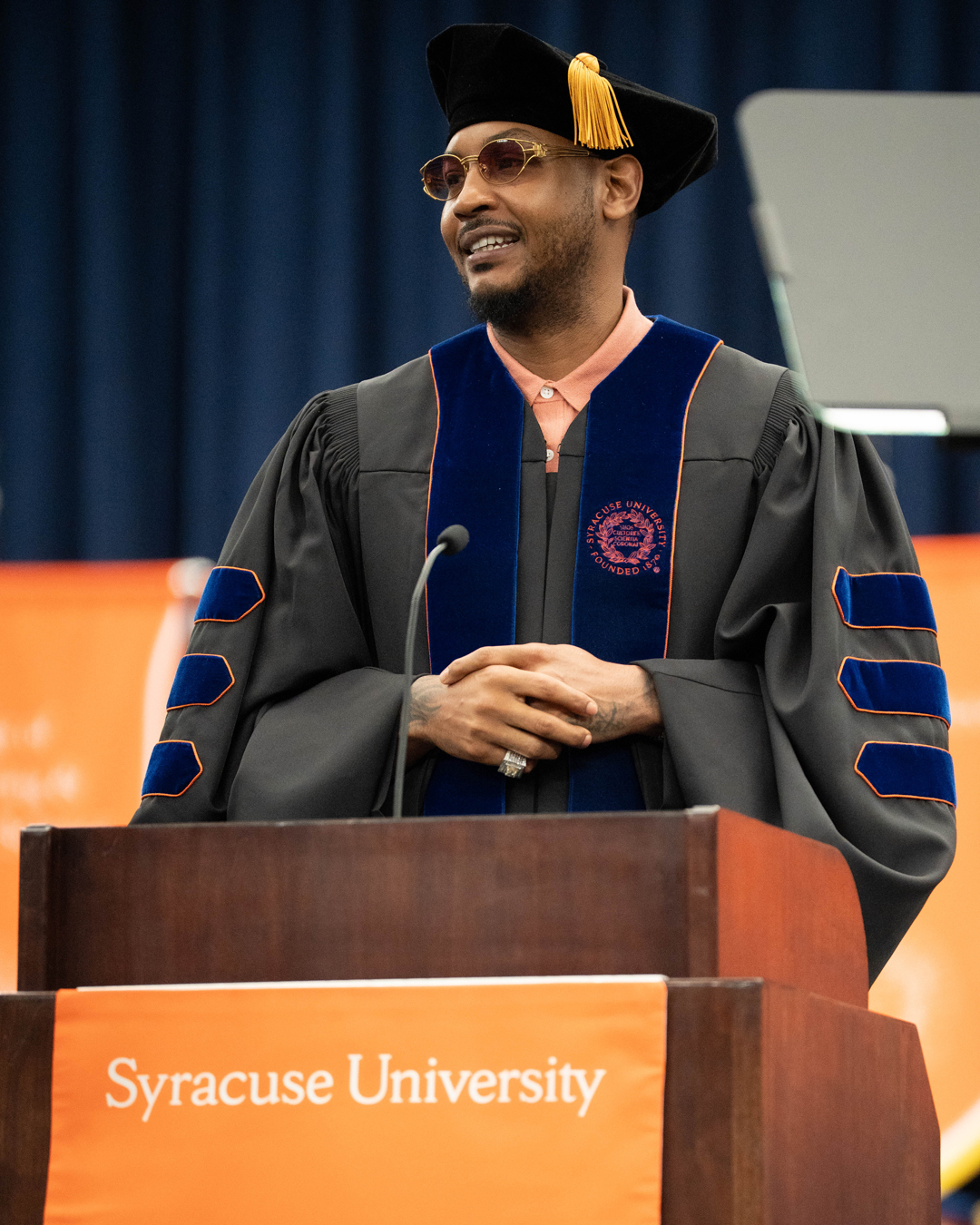 A person in academic regalia is standing at a podium with a microphone. The podium has an orange banner that reads "Syracuse University." The person is wearing a black doctoral gown with blue velvet panels and gold piping, along with a black velvet tam cap adorned with a gold tassel. The background features an orange and blue backdrop.
