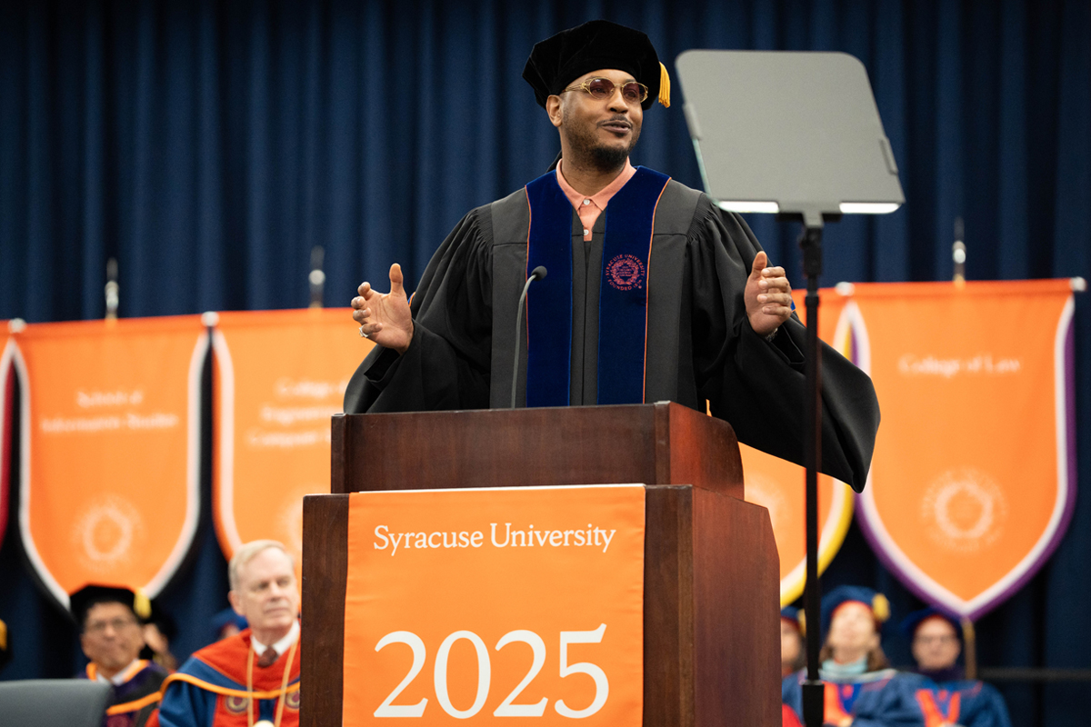 A person wearing a Commencement cap and gown and sunglasses addresses Syracuse University's graduating class of 2025 in front of a podium that reads Syracuse University 2025