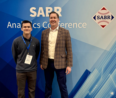 Two individuals stand in front of a blue geometric background with the text 'SABR Analytics Conference' and a logo featuring the word 'SABR' inside a baseball within a diamond shape. One person is wearing a dark sweater and name badge, and the other is wearing a plaid blazer.