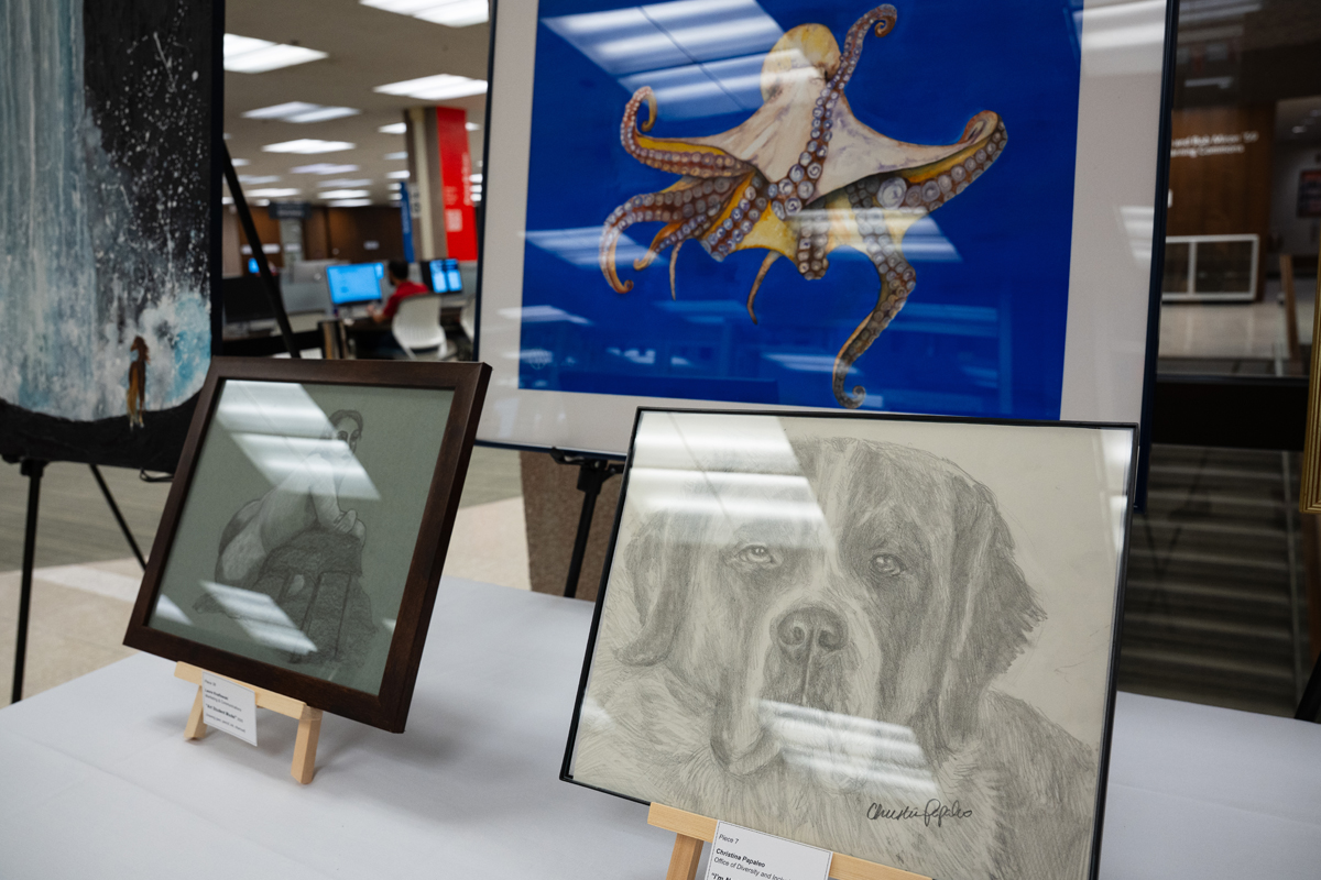 An art display featuring three framed artworks on a table. From left to right: a painting of a waterfall, a drawing of an octopus on a blue background, and a detailed pencil sketch of a dog's face signed by 'Mason Griffin.' Small placards are placed in front of each piece. The background reveals additional artworks and displays in an exhibition or gallery setting.