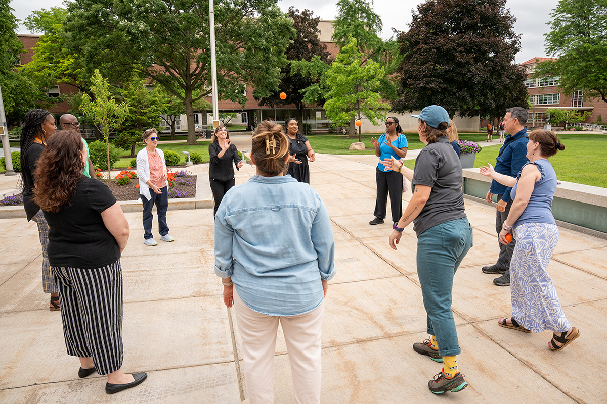 A group of people standing in a circle outdoors in a grassy courtyard, participating in a group activity. Trees and buildings are visible in the background. The individuals are casually dressed and appear to be engaged in a recreational or team-building exercise.
