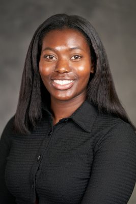 A person with long, straight hair wearing a black button-up shirt, standing against a gray background