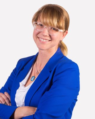 portrait of a smiling woman in a blue blazer and clear-framed glasses standing against a white backdrop with her arms crossed