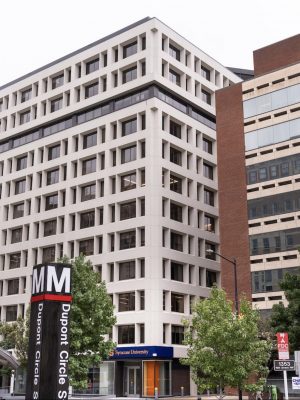 A building with a Syracuse University sign is visible behind a metro station entrance sign for Dupont Circle Station. The building has multiple windows and is surrounded by trees.