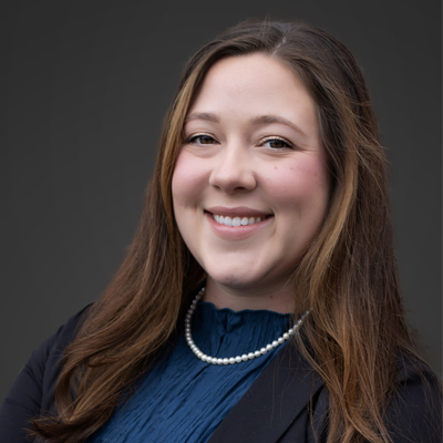 A person with long, brown hair is wearing a dark blazer over a blue top and a pearl necklace. The background is plain and dark, contrasting with the person's attire.