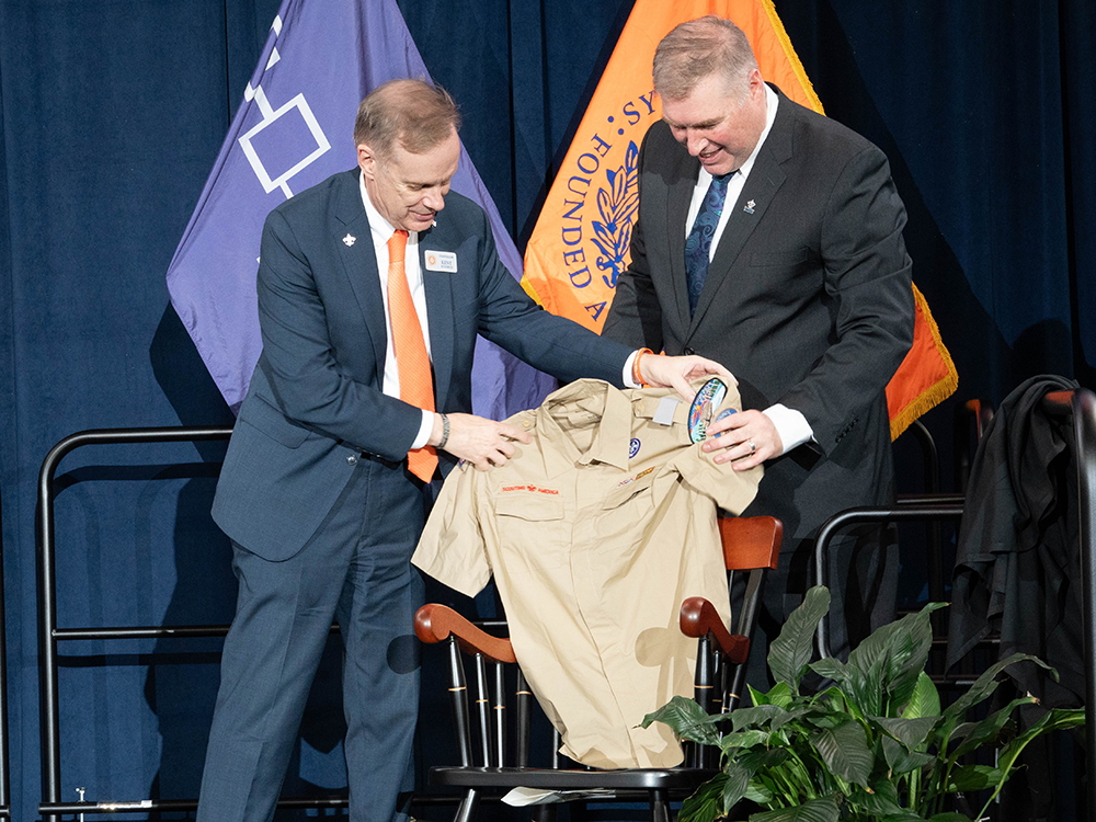 Two individuals in suits stand on a stage. One holds a beige Boy Scout uniform shirt with patches and badges, draped over the back of a wooden chair. Behind them are two flags—a Haudenosaunee flag that is purple with white symbols and an orange Syracuse University flag with the University seal. Green plants are visible in the bottom right corner.