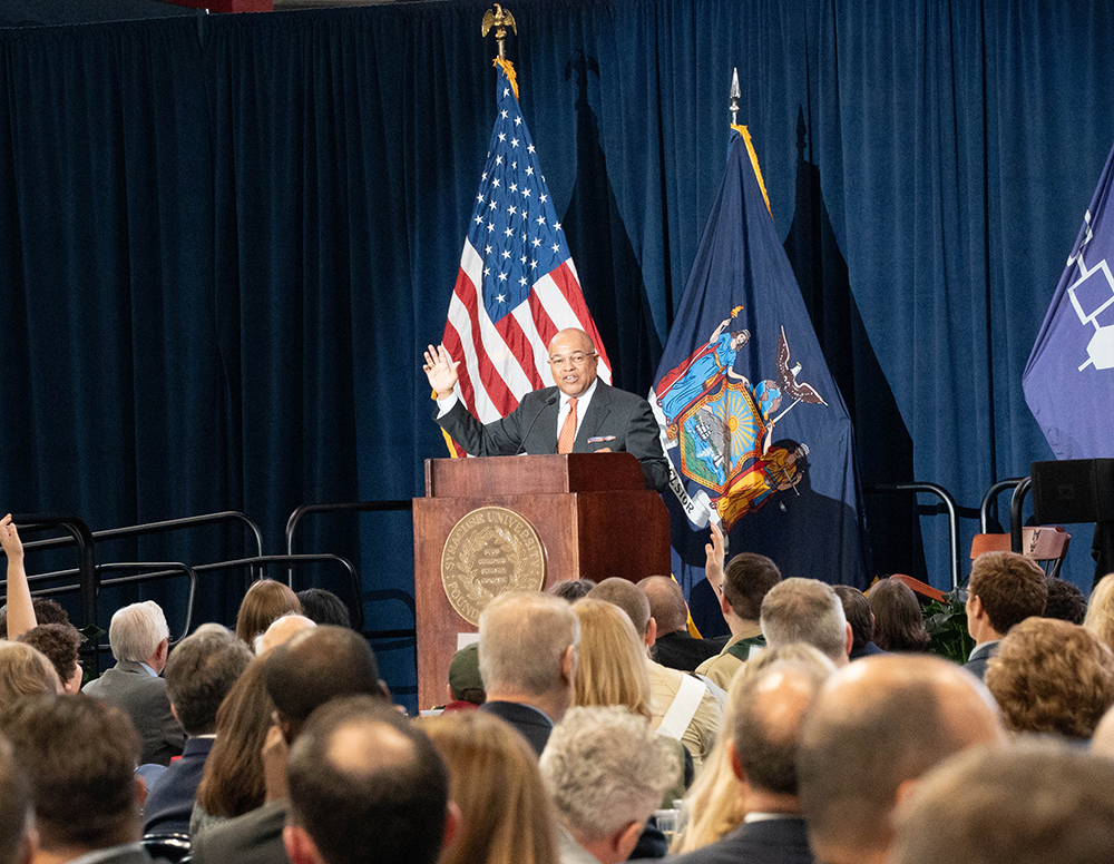 A man stands at a podium delivering a speech to an audience. The podium features a Syracuse University seal on the front. Behind the speaker are three flags: the United States flag, a state flag likely representing New York, and a purple Haudenosaunee flag. Audience members are seated and facing the speaker.