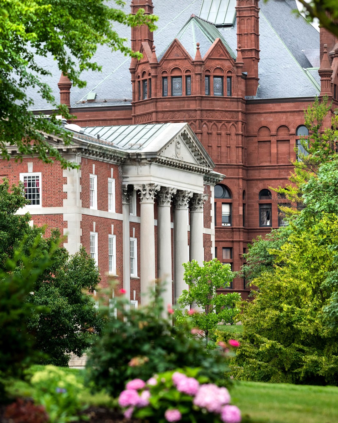 A historic brick building with tall white columns and detailed classical architecture, surrounded by trees and flowering plants in a lush, green setting.