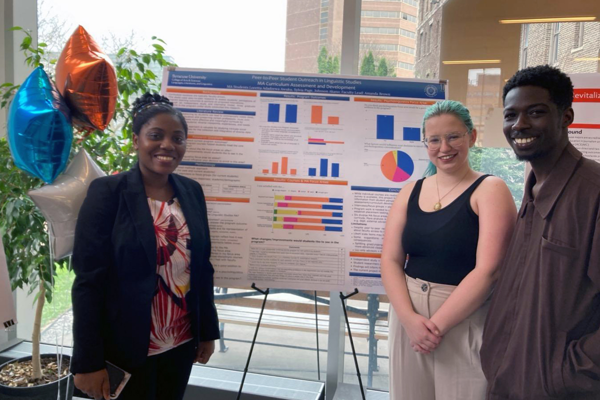 Three individuals stand in front of a scientific poster presentation, which displays various charts, graphs, and text. Behind them, a large window reveals a view of buildings and greenery.