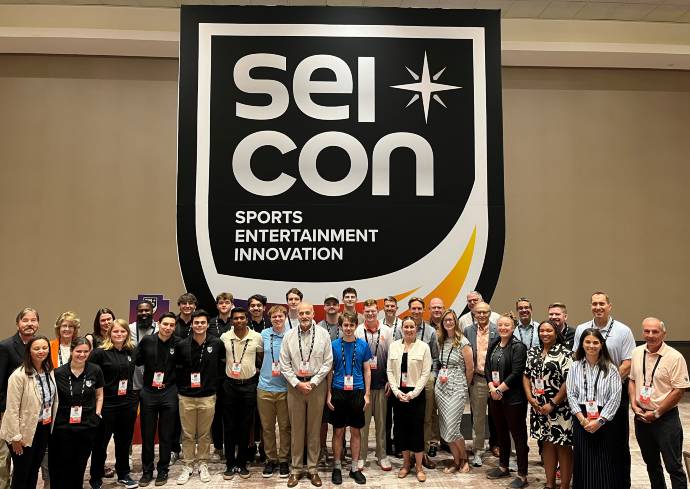 Group of people posing in front of a banner that reads "SEI CON SPORTS ENTERTAINMENT INNOVATION,' with many wearing name badges and dressed in business casual attire.