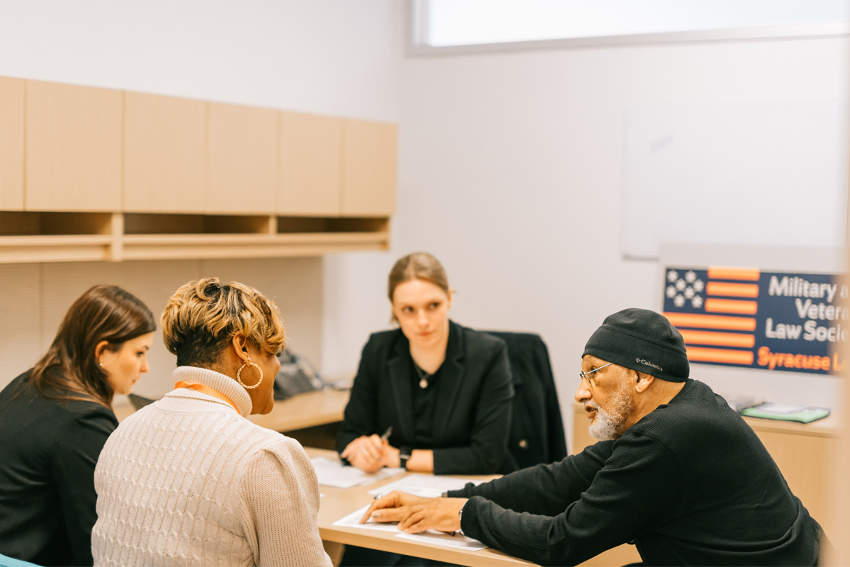 Four people are seated around a table in an office setting. The room has light-colored walls and wooden cabinets. On the right side of the image, there is a sign that reads "Military & Veterans Law Society" with an American flag graphic and "Syracuse Law" written below it.