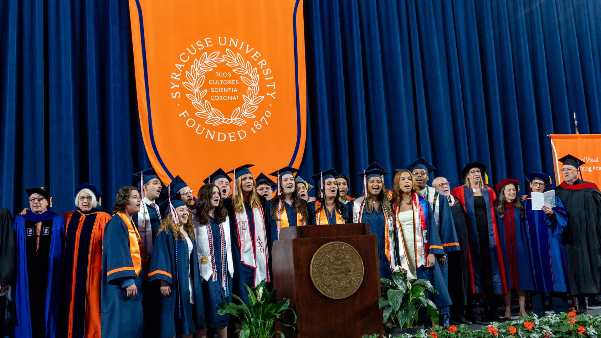 A group of people in graduation gowns and caps stand on a stage in front of a large orange banner with the text "Syracuse University Founded 1870" and a laurel wreath design. The stage is decorated with plants, and there is a podium with the university seal in the center. Some individuals are holding diplomas, and others are wearing academic regalia
