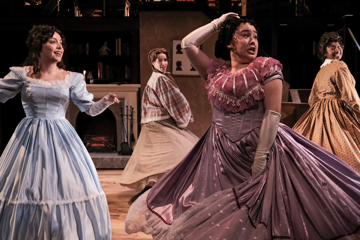 Four individuals dressed in 19th-century period costumes perform a dance or theatrical scene on a wooden stage. The backdrop features bookshelves and a fireplace. The performers wear long dresses in light blue, purple, plaid with beige, and mustard yellow.