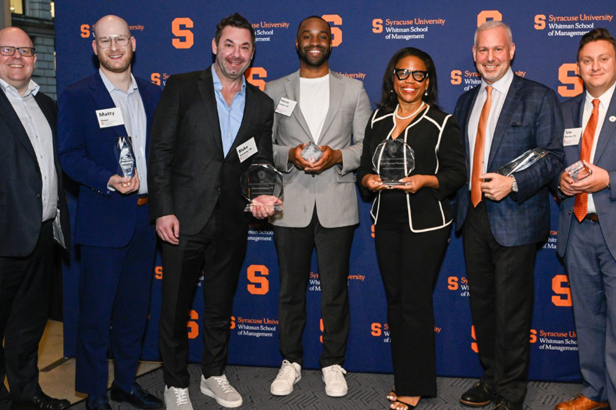 Seven individuals in business attire stand in front of a backdrop featuring the Syracuse University Whitman School of Management logo. Each person is holding an award.