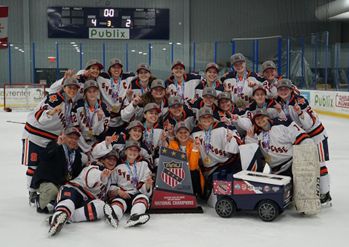 The image shows a group of ice hockey players posing together on an ice rink. They are wearing matching uniforms with "Syracuse" written on them and are celebrating a victory. The scoreboard in the background shows a final score of 4-2. The team is holding a large trophy that reads "AAU National Champions." There is also a small cart with equipment next to the team.