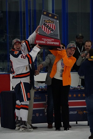 The image shows a group of people on an ice rink, with one person in a hockey uniform and another person in a suit holding up a large trophy that reads "AAU NATIONAL CHAMPIONS." The background includes other individuals and some banners.