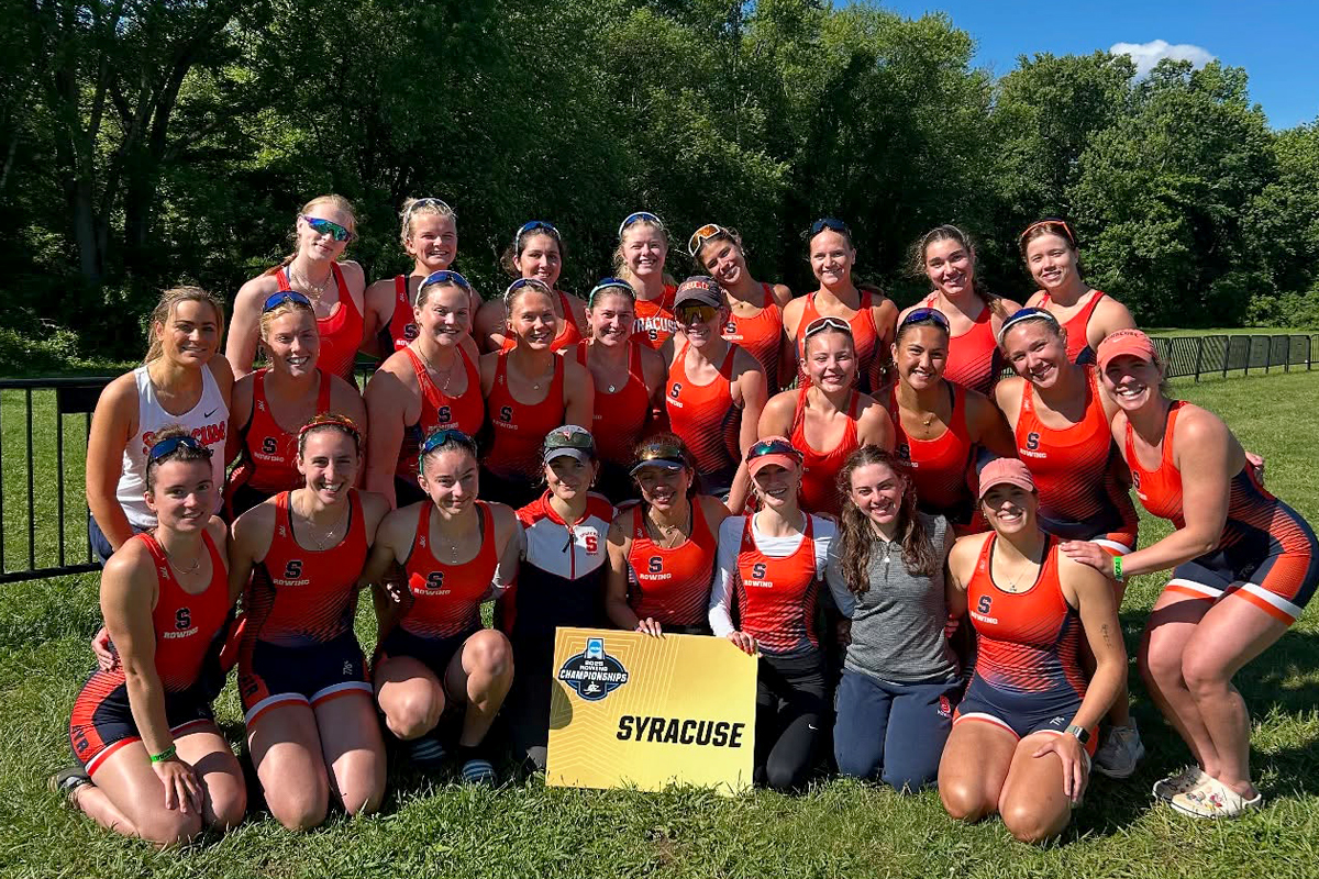 A group of athletes in orange and blue uniforms with the letter 'S' pose outdoors in front of a wooded area, holding a yellow sign that reads 'NCAA Championships SYRACUSE.