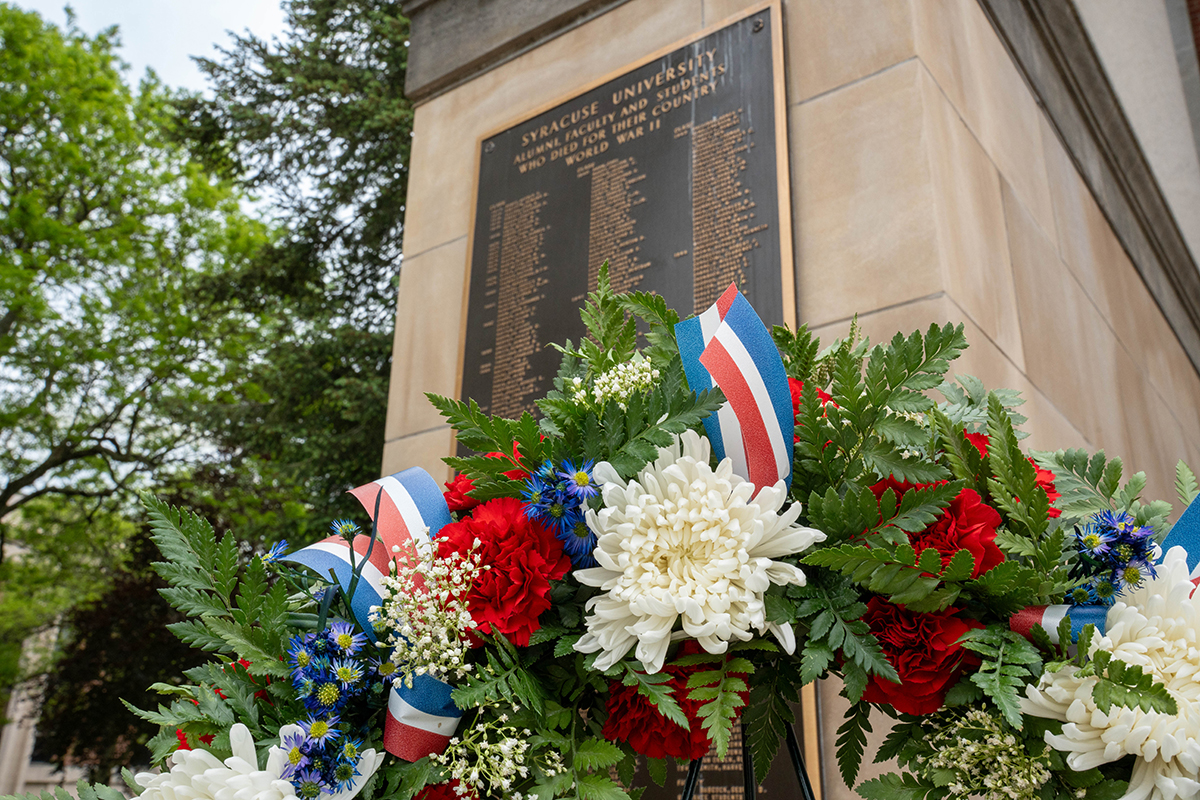 A memorial plaque with names inscribed on it is mounted on a stone structure. In front of the plaque is a wreath made of red, white, and blue flowers with green foliage and matching ribbons. Trees and part of a building are visible in the background.