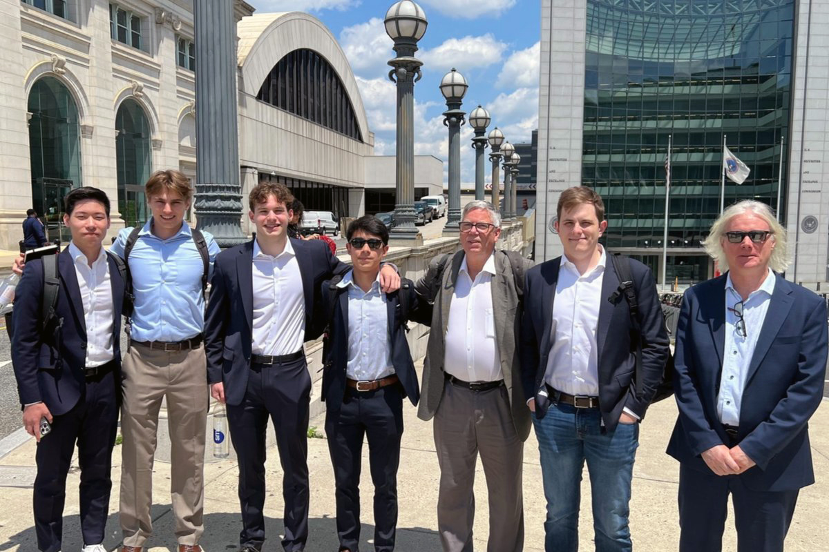 Seven men dressed in business attire, including suits, dress shirts, and ties, standing outdoors in front of a building with columns and a large arched window. The background includes a clear blue sky with scattered clouds, street lamps, and a nearby building with glass windows.