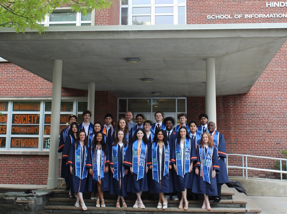 Graduates in caps and gowns standing on the steps of Hinds Hall, with words like 'Social', 'Cybersecurity', and 'Networks' displayed on the building's windows
