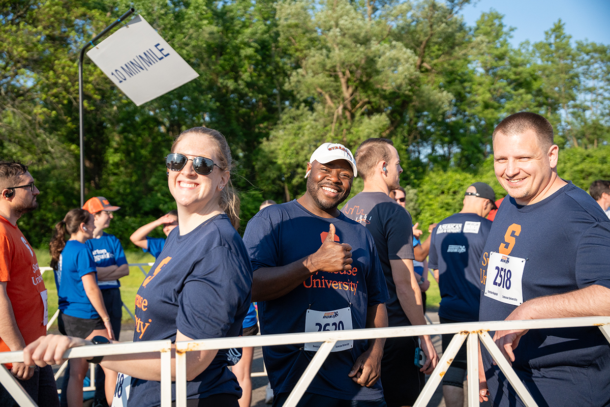 A group of people participate in the Syracuse WorkForce Run. wearing 'Syracuse University' shirts and numbered bibs. A sign in the background reads '10 MIN/MILE,' and the setting includes trees and greenery.