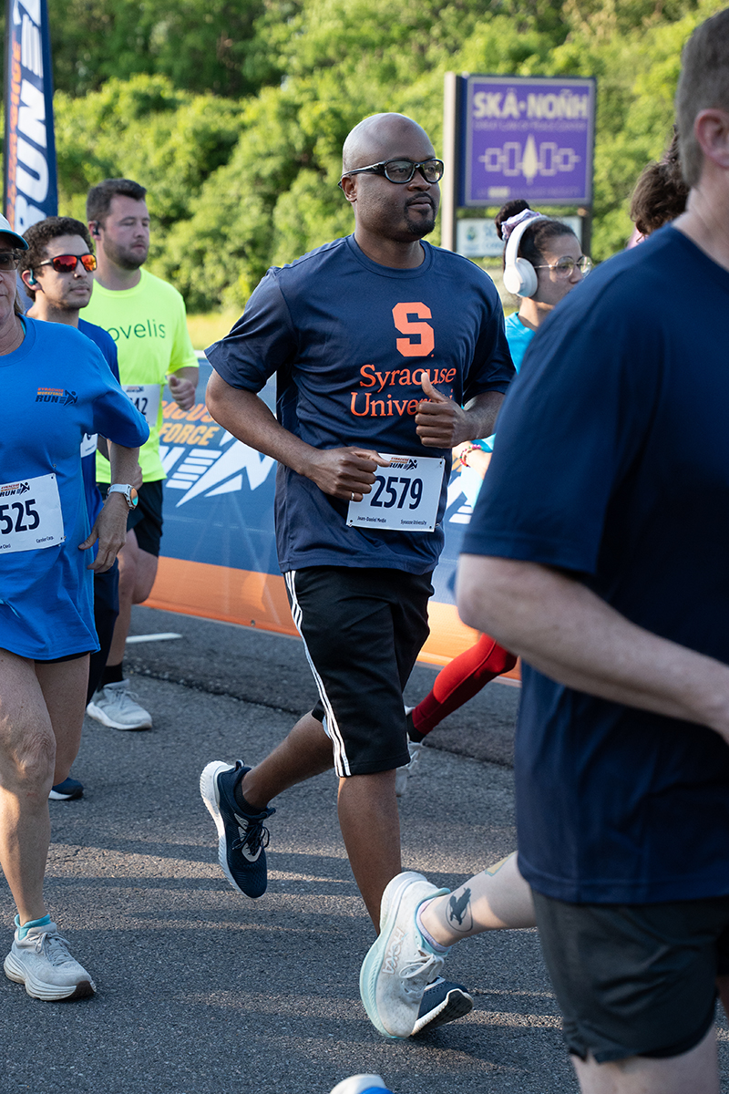 A group of people participate in a running event. The central figure wears a navy blue Syracuse University t-shirt and a race bib numbered 2579.