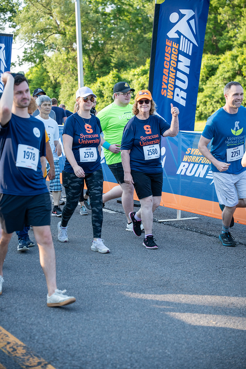 A group of people participate in the Syracuse WorkForce Run, wearing athletic clothing and numbered bibs including 1978, 2519, 2580, and 2751. Banners in the background read 'SYRACUSE WORKFORCE RUN,' and some participants wear shirts with 'Syracuse University' printed on them.