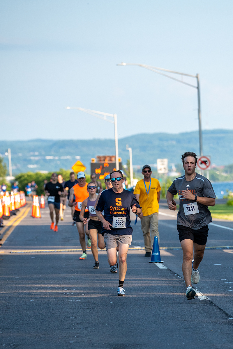 A group of people run in a race on a road, with several runners wearing numbered bibs. The runner in the foreground on the right has bib number 3241, and another nearby wears a Syracuse University shirt with bib number 2638. Traffic cones, race signs, and a scenic backdrop of hills or mountains are visible.