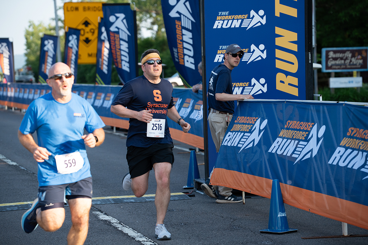 Two runners participate in a race. The runner on the left wears a blue shirt and black shorts with bib number 559. The runner on the right wears a navy blue Syracuse University shirt, black shorts, and bib number 2516. Banners along the route read 'Syracuse Workforce Run'