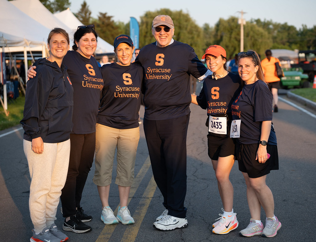 Six people stand together on a road wearing navy blue shirts with 'Syracuse University' and an orange 'S' logo. Two individuals have race bibs numbered 1435 and 1451. Tents and other participants are visible in the background.