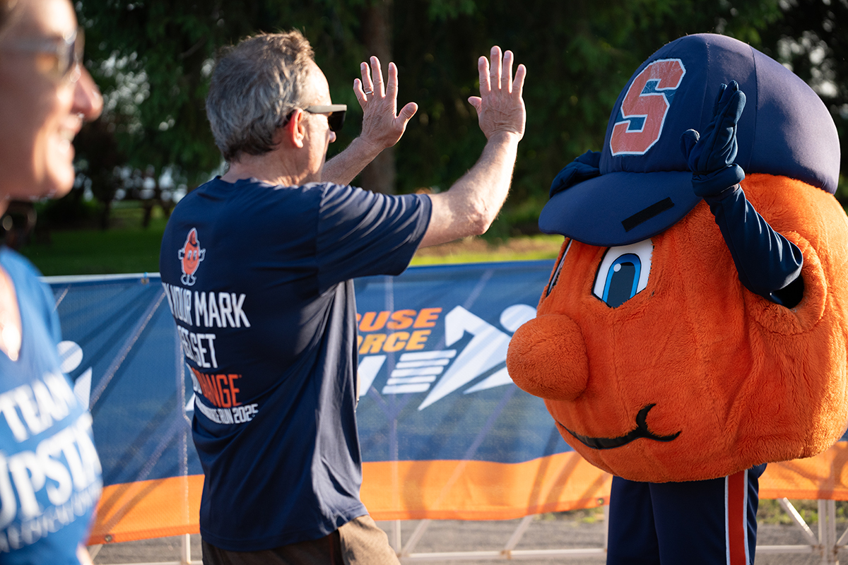 A man gives a high-five to Syracuse University mascot Otto the Orange. The man wears a navy blue shirt with the text 'ON YOUR MARK GET SET. GO ORANGE' on the back.