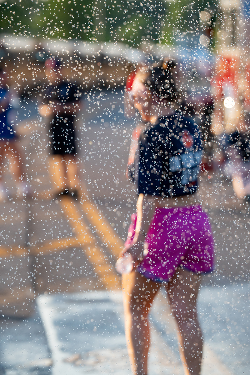 A group of people outdoors, with one person in the foreground wearing a dark blue shirt and pink shorts. The image is captured through a spray of water droplets, creating a speckled effect. The background is slightly blurred with other individuals visible.