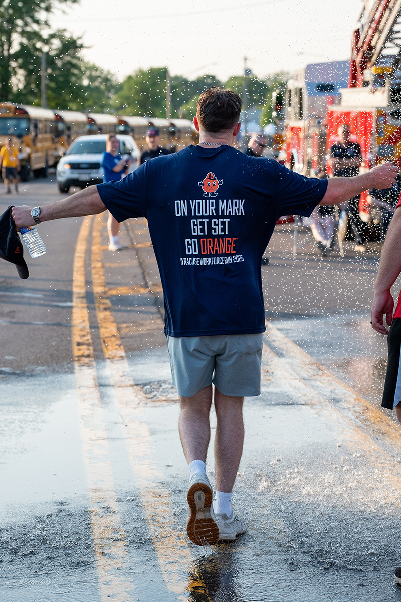 A person wearing a navy blue shirt and gray shorts walks down a wet street with arms outstretched. The back of the shirt reads 'ON YOUR MARK GET SET GO ORANGE' with 'SYRACUSE WORKFORCE RUN 2025' below. The person holds a water bottle, with water droplets spraying around. School buses, emergency vehicles, and other participants are visible in the background.