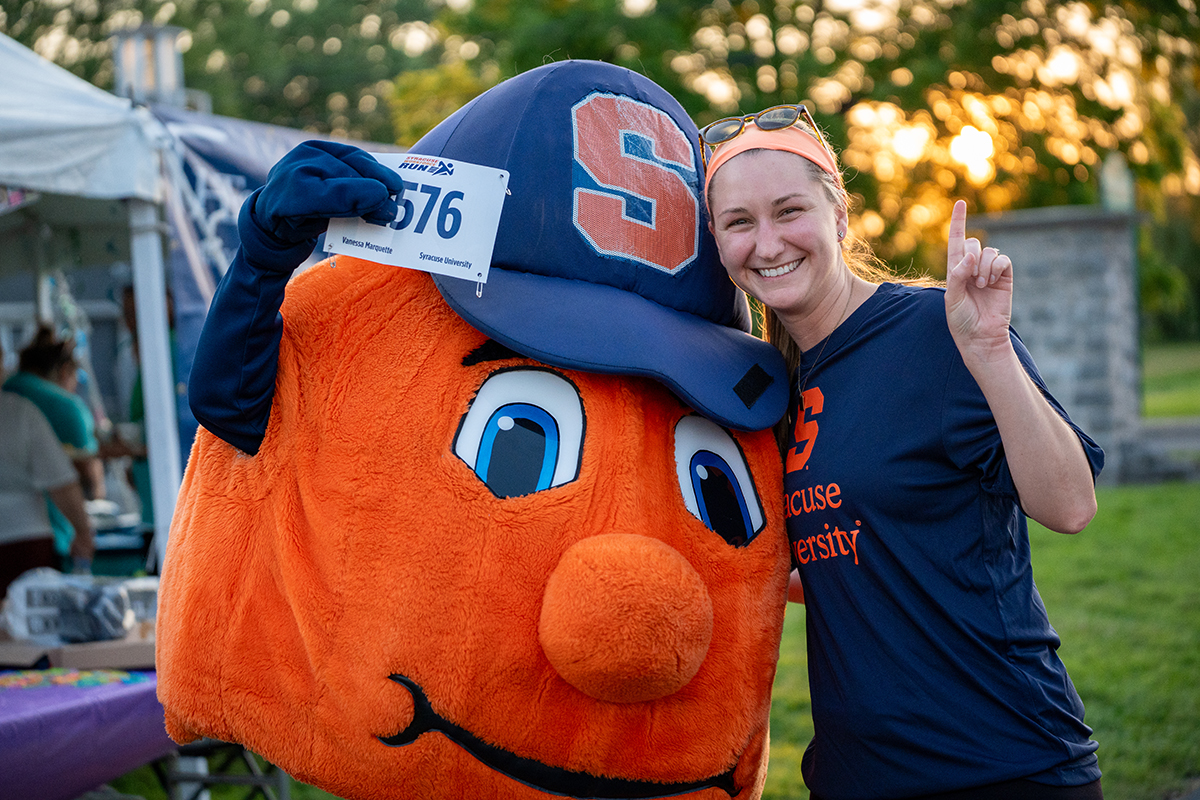 A person poses with the Syracuse University mascot, Otto the Orange, who is wearing a large blue hat with an orange 'S' and a race bib numbered 576. The person wears a navy blue 'Syracuse University' shirt and holds up one finger. Tents and trees are visible in the background.