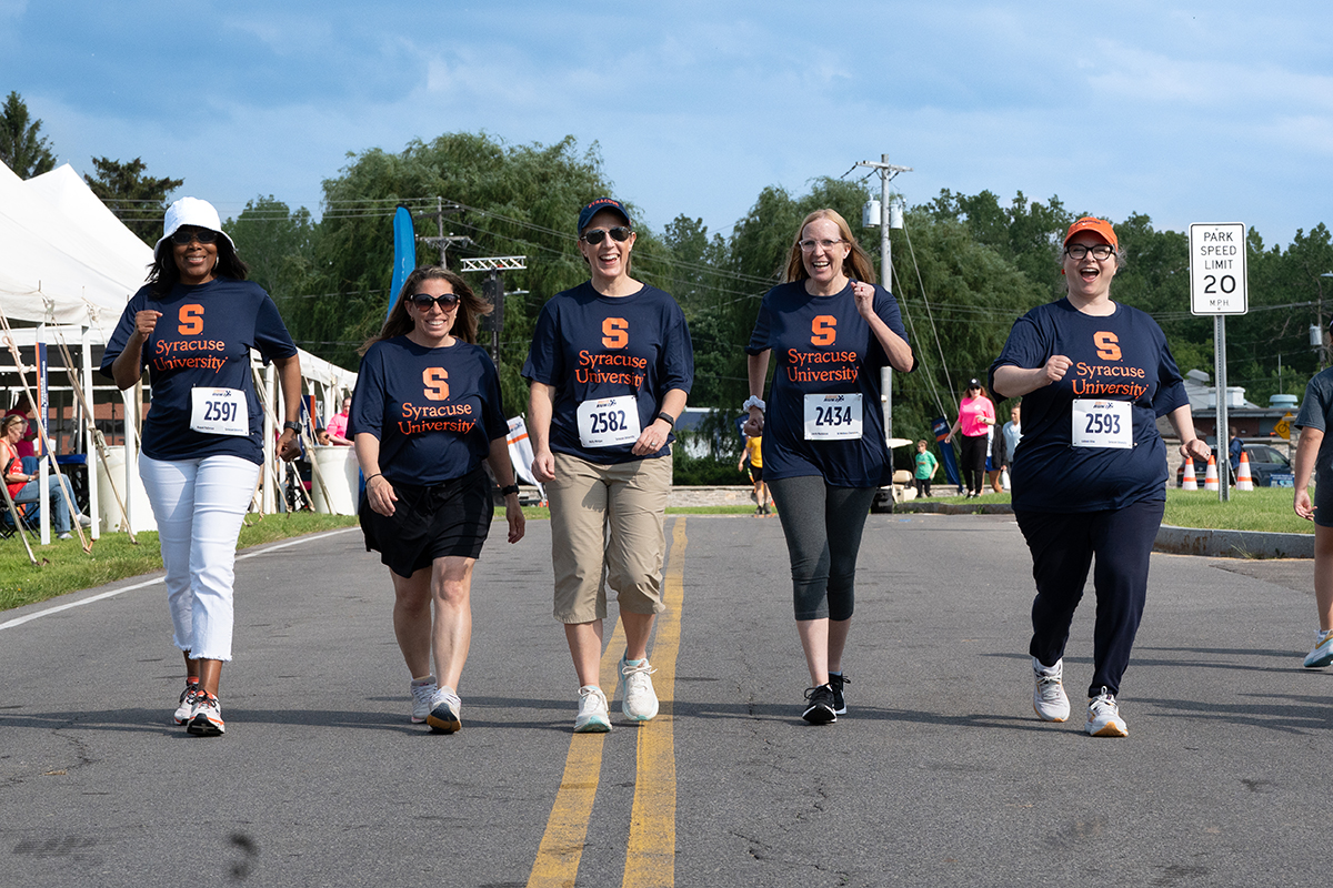 Five individuals walk along a road during a race or walk event, wearing navy blue t-shirts with an orange 'S' and 'Syracuse University' text. Each has a race bib with numbers including 2597, 2582, 2434, and 2593.