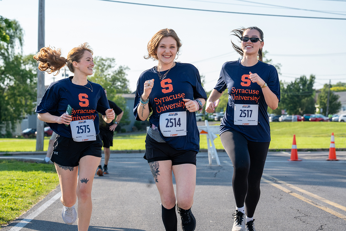 Three individuals run on a road wearing navy blue shirts with an orange 'S' and 'Syracuse University' text. Race bibs numbered 2515, 2514, and 2517 are visible. The background features trees, greenery, parked cars, and traffic cones.