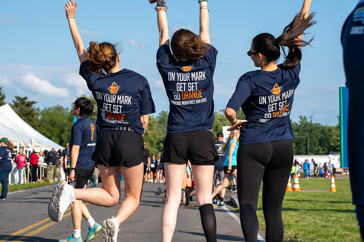 Three people seen from behind wearing matching navy blue shirts with the text: 'ON YOUR MARK GET SET GO ORANGE SYRACUSE WORKFORCE RUN 2025.' They are outdoors at a race, with other participants and spectators in the background.